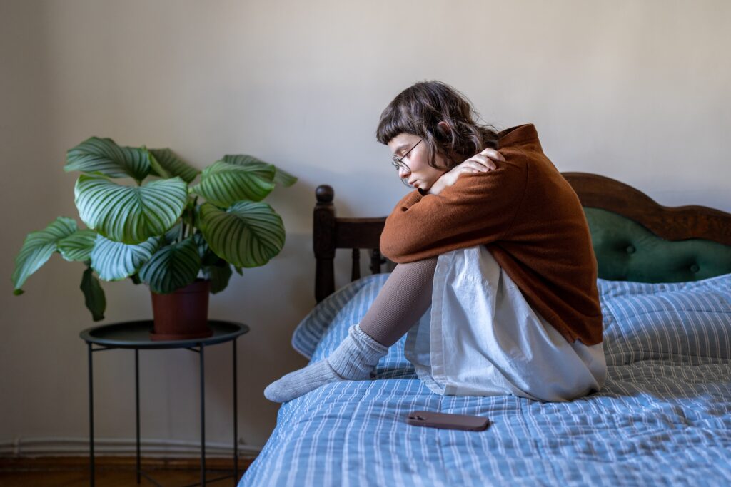 Gefahren im Netz: Traurige Teenagerin sitzt zusammengekauert auf ihrem Bett mit Smartphone neben sich