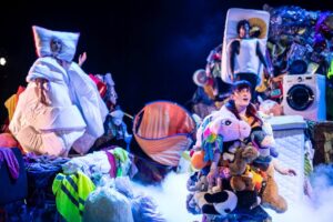 Junge Triennale 2026 – Szene aus „Waste Me“ mit Performerinnen und Performern zwischen Kleidung, Spielzeug und Alltagsgegenständen